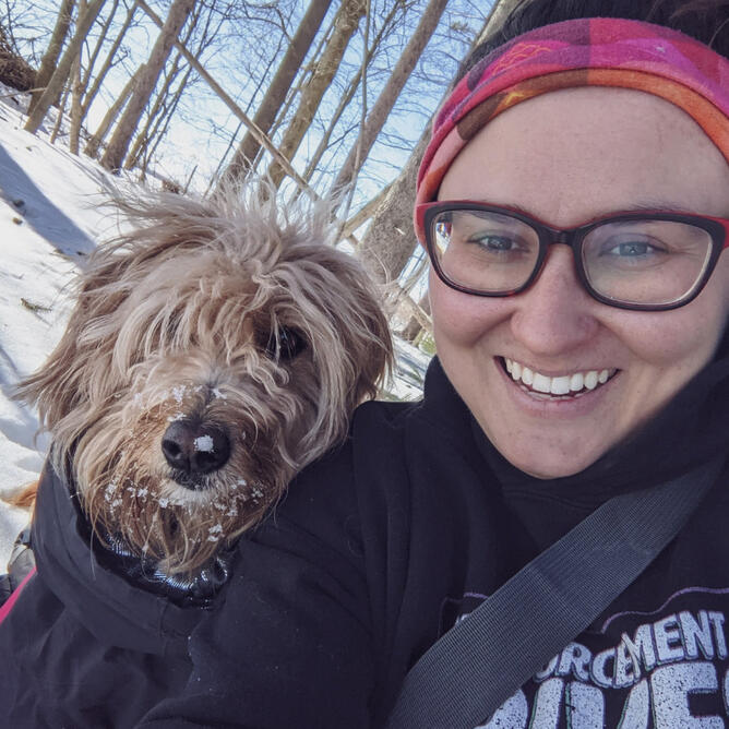Klarissa & Chelsea Klarissa is a femme non-binary person with brown hair, Chelsea is a blonde mini doodle, they are in a snowy forest.