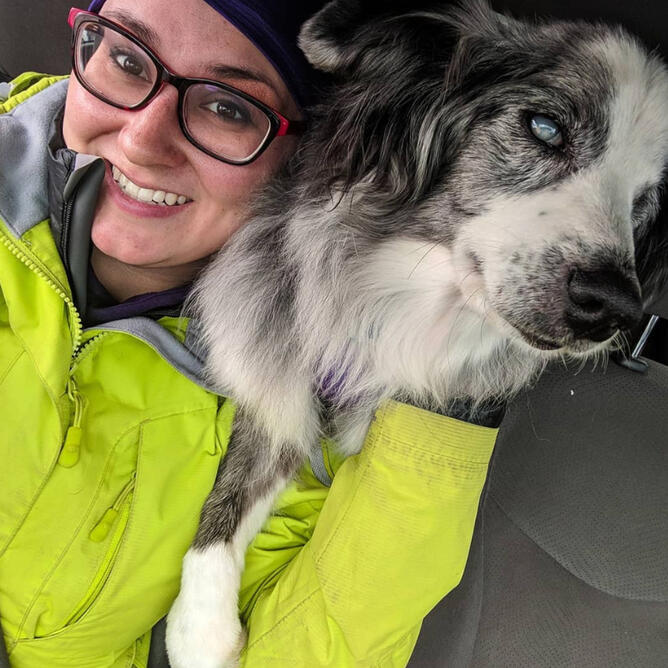 Klarissa & Ellie Klarissa is a femme non-binary person with brown hair & glasses. They are wearing a lime green rain coat, while Ellie, a blue merle mini Aussie stands over their shoulder.