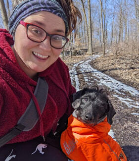Klarissa I am a white, femme non-binary person with brown hair & brown eyes. I wear black, plastic framed glasses & hair pulled into a messy bun. I am in a red fleece sweater, squatting down to take a selfie with Dyson. He is wearing a bright orange winter coat, be
